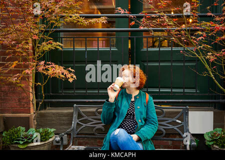 Young redhead woman drinking coffee on bench in front of nice cafe. Stock Photo