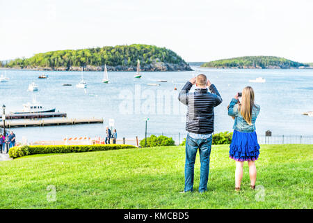 Bar Harbor, USA - June 8, 2017: Young couple, man, woman taking pictures using smartphone, mobile phone on green grass hill in park downtown village i Stock Photo