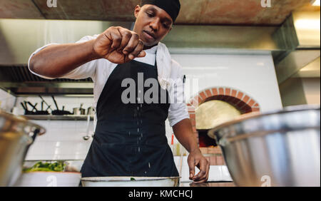 Chef sprinkling spices on dish in commercial kitchen. African male cook preparing food in restaurant kitchen, adding seasoning. Stock Photo
