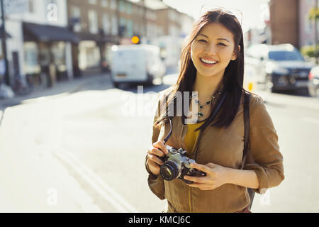 Portrait smiling, confident young female tourist with camera on sunny urban street Stock Photo