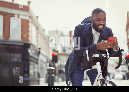 Young businessman commuting with bicycle, texting with cell phone on sunny urban street Stock Photo