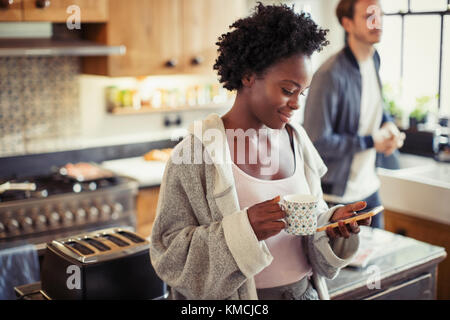 Woman drinking coffee, texting with smart phone in kitchen Stock Photo