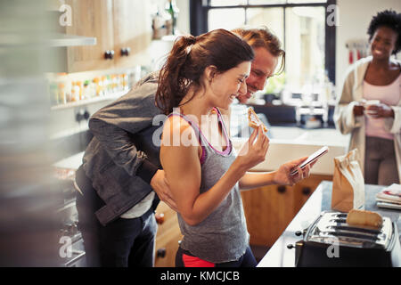 Smiling couple texting with smart phone, eating toast in kitchen Stock Photo
