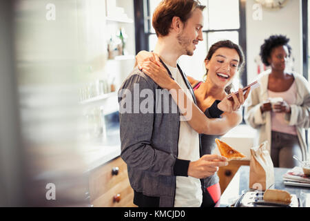 Affectionate couple hugging, texting with smart phone in kitchen Stock Photo