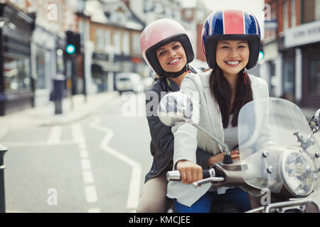 Smiling young women friends wearing helmets, riding motor scooter on urban street Stock Photo