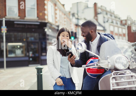 Young couple laughing, using cell phone at motor scooter on sunny urban street Stock Photo