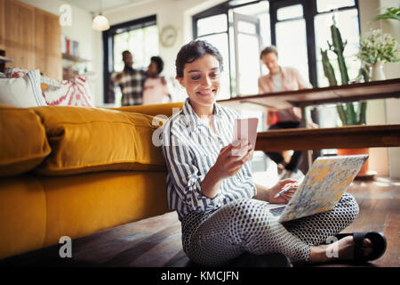 Smiling young woman using laptop and texting with smart phone on living room floor Stock Photo