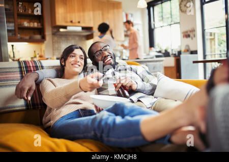 Smiling young couple watching TV, drinking beer and eating popcorn on living room sofa Stock Photo