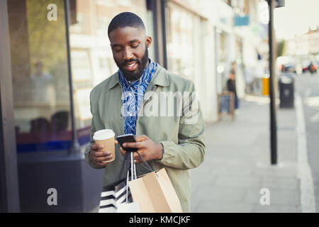 Young man with coffee and shopping bags texting with cell phone on urban sidewalk Stock Photo