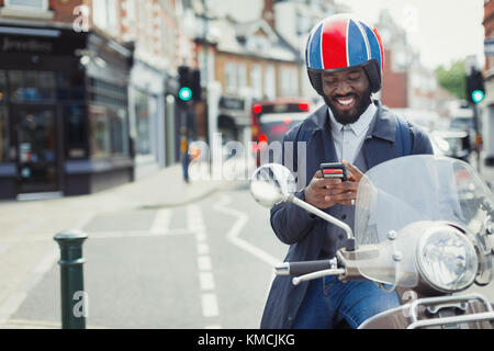 Smiling young businessman in helmet on motor scooter texting with cell phone on urban street Stock Photo