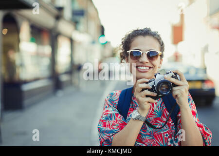 Portrait smiling young female tourist in sunglasses photographing with camera on urban street Stock Photo