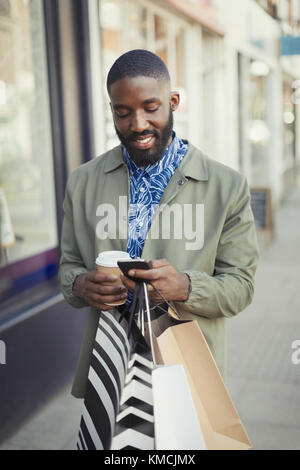 Smiling young man with coffee and shopping bags texting with cell phone on urban sidewalk Stock Photo
