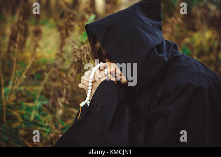 a monk in a black robe praying in the woods on his knees Stock Photo