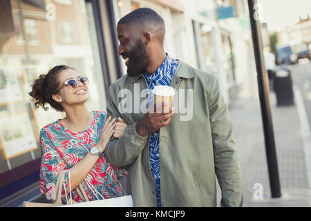 Smiling young couple with coffee walking arm in arm along storefronts Stock Photo
