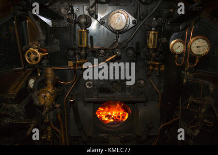 Night shot inside cab of steam locomotive, looking into the lit fire chamber of the boiler backplate. Valves, rods, pipes, pressure gauges all visible Stock Photo