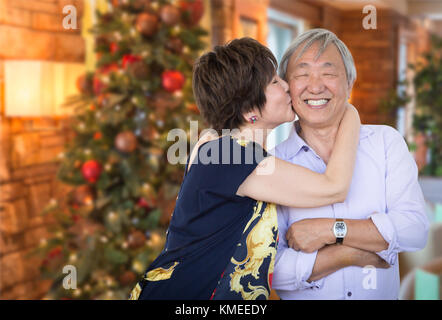 Happy Senior Chinese Couple Kissing In Front of Decorated Christmas Tree. Stock Photo