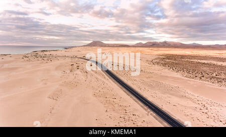 Aerial view of road through sandy desert dunes towards sea and volcanic mountains, sunset sky, Corralejo, Fuerteventura, Canary Island, Spain Stock Photo