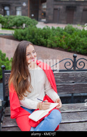 An attractive girl with long brown hair and a white tooth smile sits on a bench and writes her thoughts on the urban background in a red notebook. She Stock Photo