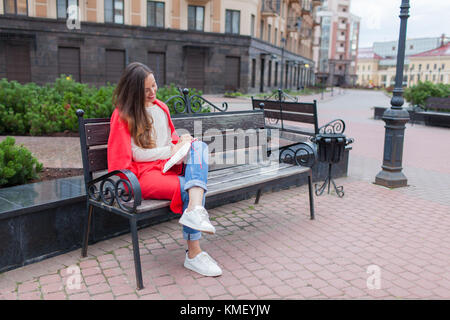 An attractive girl with long brown hair and a white tooth smile sits on a bench and writes her thoughts on the urban background in a red notebook. She Stock Photo