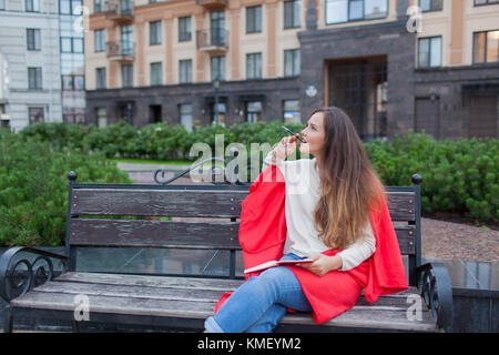 An attractive girl with long brown hair sits on a bench, hiding behind a red rug, gnawing a pen and thinking on an urban background. She has a noteboo Stock Photo