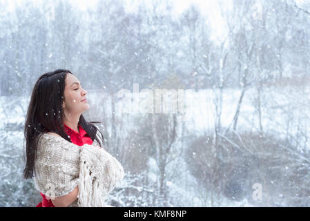 Attractive young woman with long dark hair, in a red dress, covered by a handmade shawl, with her eyes closed, enjoying the snowfall. Stock Photo