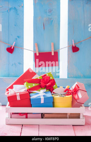 Colorful gift boxes in a white wooden crate on a pink table with a red paper note and hearts tied to a string, on a blue fence. Stock Photo