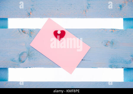 Blank pink paper note with a red heart glued to it and together pinned to a blue wooden fence. Stock Photo