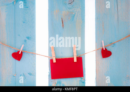 Empty red message card surrounded by red hearts, tied to a linen string with wooden clips, on a blue fence. Stock Photo