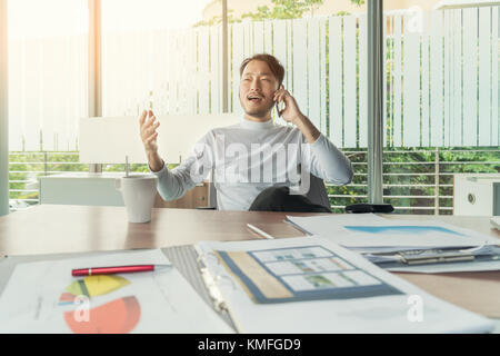 Portrait of Asian smiling businessman or architect speaking on mobile phone, sitting at desk. Stock Photo