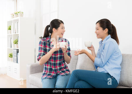 leisurely pretty girls sitting on living room sofa talking together and drinking hot coffee chatting enjoying afternoon tea time. Stock Photo