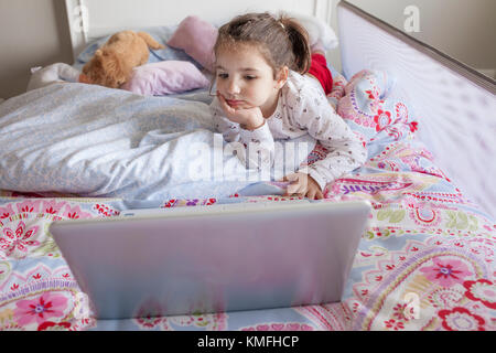 Little girl lying in bed and watching cartoons with a laptop in his bedroom. She looks entertained Stock Photo