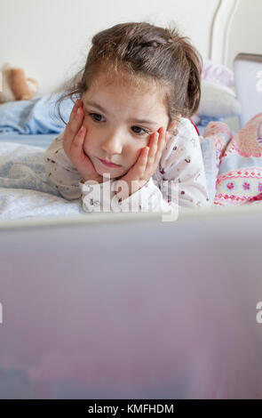 Little girl lying in bed and playing with a laptop in his bedroom. She looks inattentive Stock Photo