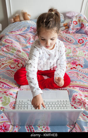 Little girl sitting in bed and playing online games with laptop computer. She is touching the screen Stock Photo