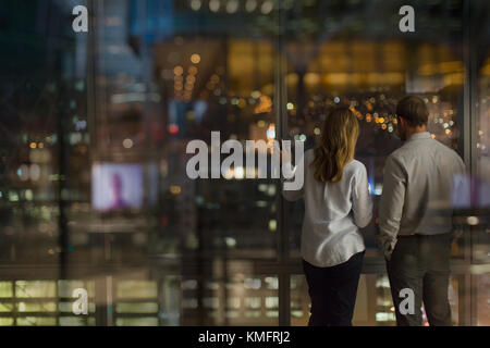 Businessman and businesswoman looking out urban office window at night Stock Photo