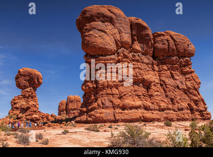 A group of tourists show the massive scale of the eroded sandstone formations in Arches National Park. Balanced Rock is on the left, and is 128ft (39m Stock Photo