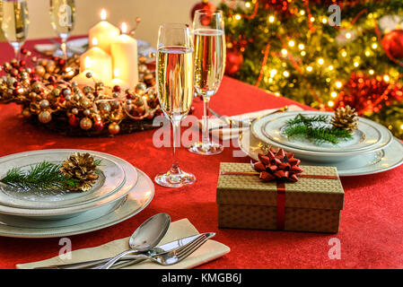 Beautifully decorated Christmas table - gift, glasses of champagne and tableware with Christmas decoration on a red tablecloth. Stock Photo