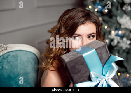 Attractive woman holding a Christmas gift tied with a blue bow to her face as she sits in a matching blue vintage chair in front of a decorated tree. Stock Photo