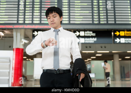 Asian young businessman looking at his watch in front of airport timetable at the airport terminal. Business travel concept. Stock Photo