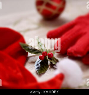 closeup of a santa hat, a pair of cozy red gloves, a red chrismtas ball and a twig of holly on a white rustic  wooden surface Stock Photo