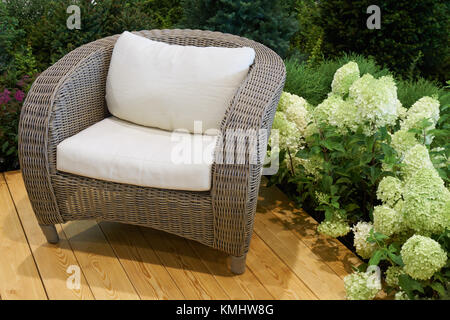Resting place in the garden surrounded by coniferous trees and flowers. Wicker chair with comfortable pillows on wooden deck Stock Photo