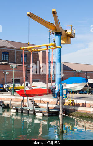 Giudecca boatyard, Venice, Italy. Red boat with outboard motor suspended midair in slings on a boat lift or crane above the quay during launching. The Stock Photo