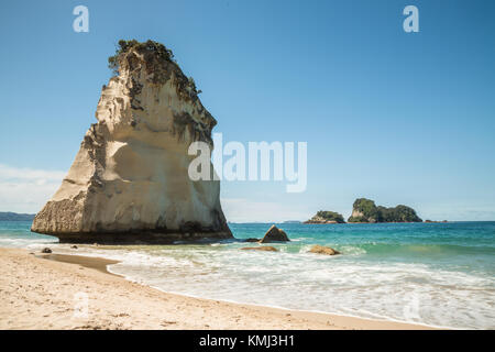 A sea stack off the coast of the Coromandel Penninsula stands above breaking waves on a sandy beach. Above the blue-green waters of the Pacific Ocean  Stock Photo