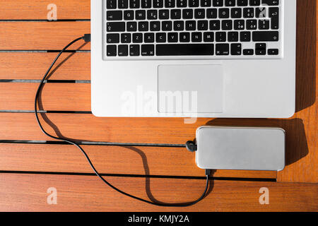 White smart phone charges and is connected by usb cable to metallic laptop with black keyboard on orange wooden table outside in cafe or restaurant. Stock Photo