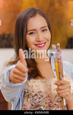 Beautiful young woman painter with paint brush in hands and pointing in from of her a thumbs up, in a blurred background Stock Photo