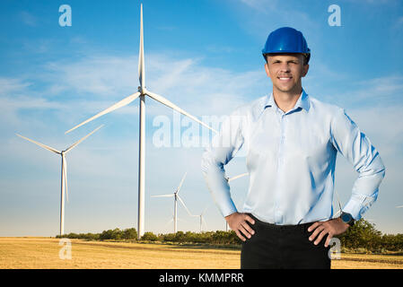 Young blue collar worker or engineer wearing in yellow helmet stands in the field with wide smile against wind turbines farm. Half-length portrait. Stock Photo