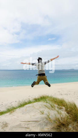 Young model jumping on a sand dune with open arms. White sandy beach and blue sky in the background. Scotland, UK. Stock Photo