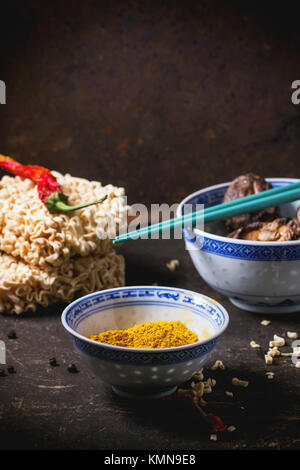 Dry noodles for ramen soup with tumeric powder and shiitake mushrooms in porcelain bowls, served with red hot chili peppers over dark table. See serie Stock Photo