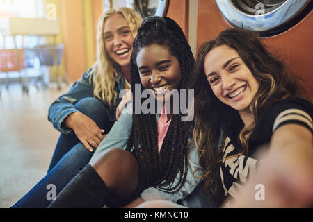 Group of smiling young female friends sitting on a laundromat floor taking selfies while doing laundry together Stock Photo