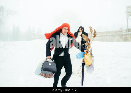 The cheerful groom with the knitted scarf on his head is holding the things for the winter picnic while his beautiful lover is standing behind. Stock Photo