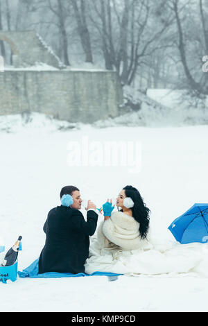 Full-lenght back view of the beautiful couple of newlyweds drinking champagne while sitting on the plaid during their blue winter picnic on the snowy meadow. Stock Photo